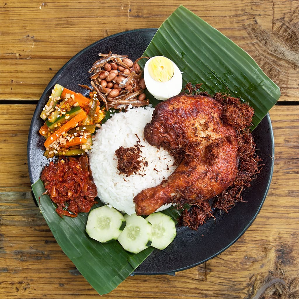 Malaysian Nasi Lemak plate with fragrant rice, spicy sambal, crispy ikan bilis (anchovies) and peanuts, fresh cucumber slices, hard-boiled egg, and golden fried chicken – a traditional Malay breakfast delicacy.
